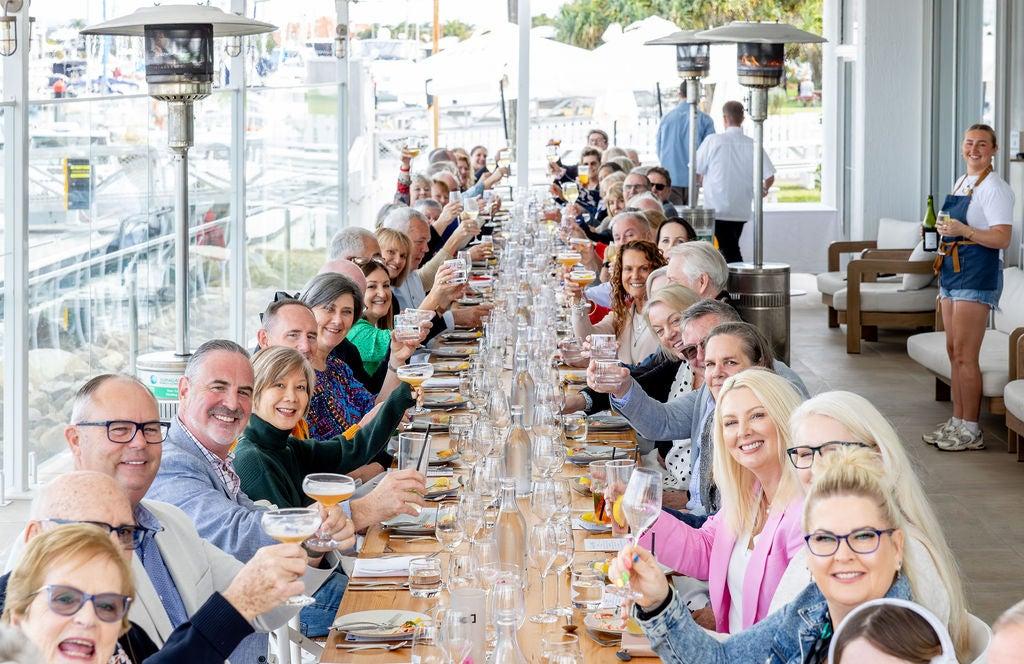 A long table setting with guests holding up glasses to cheers to the camera.