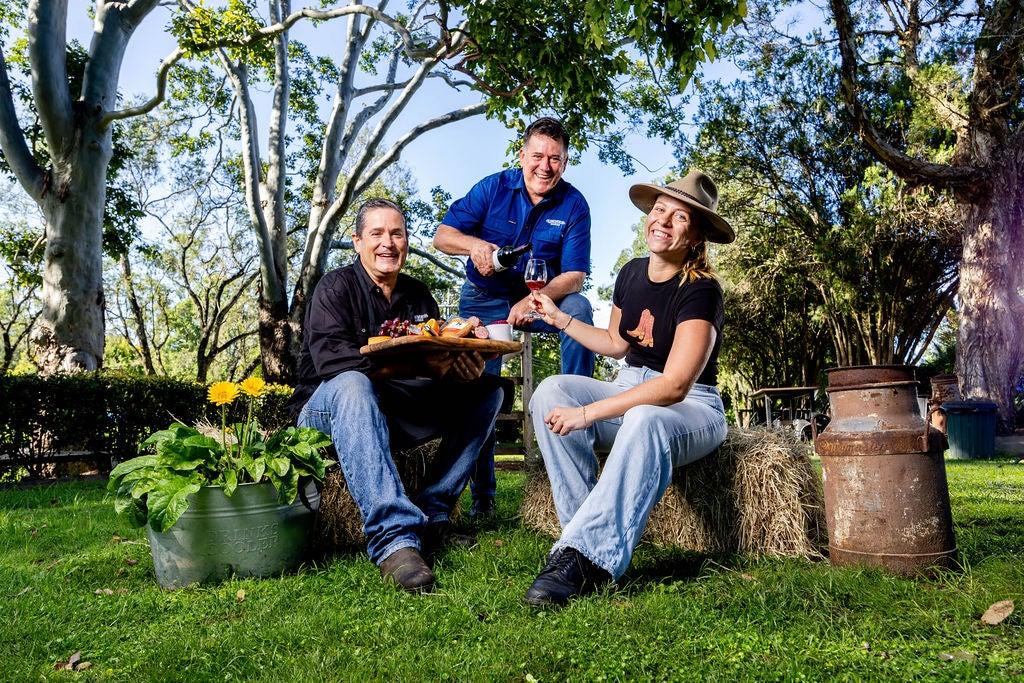 Paul Campbell and Carlotta Biemans sitting on hay bales, with Dominc Ovenden leaning in pouring wine into the glass Carlotta is holding.