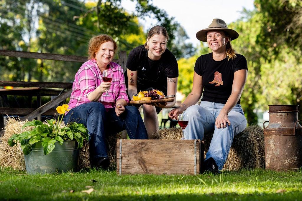 Lisa O-Farrell (in a pink shirt) holding a glass of wine and Carlotta Biemans (wearing a hat) sitting on hay bales, and Ally Richter leaning in presenting a food platter.