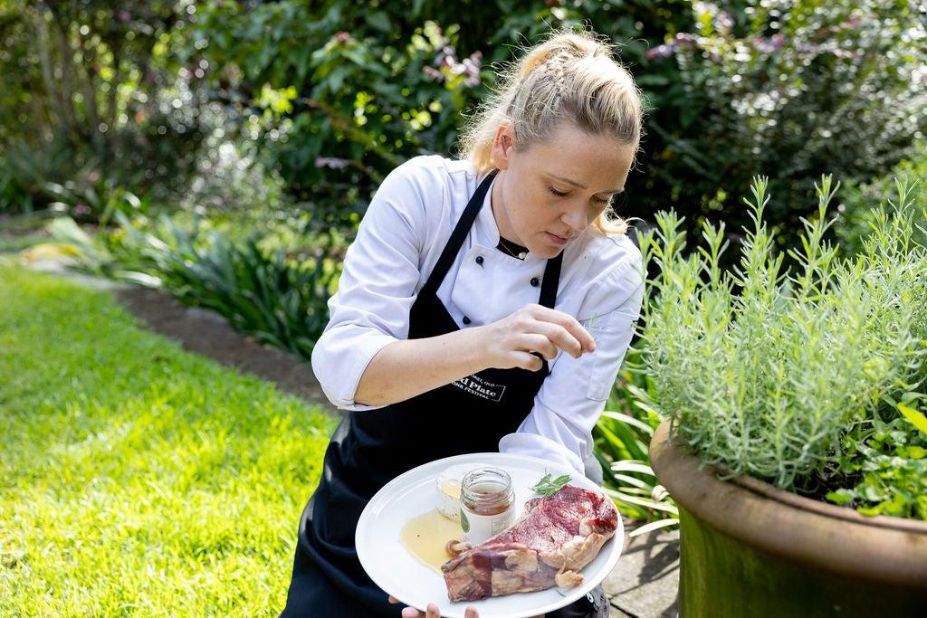 Chef Sophie Phipps holding a plate of food - meet and jar of honey - her hand is lifted as if she is dropping a pinch of salt or spice on the plate. A beautiful green garden is beside and behind her.