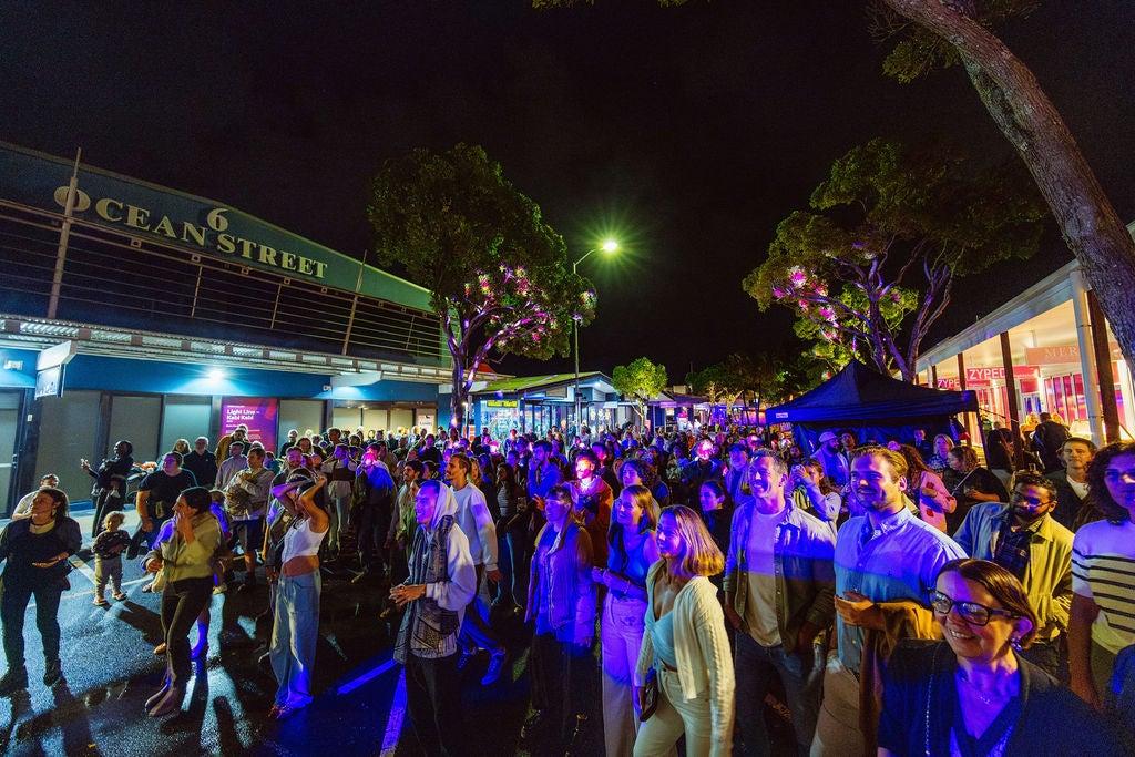 Taken from the side of stage, looking towards a crowd of people, some bathed in purple light. Gathered in Ocean Street to watch a Horizon performance.