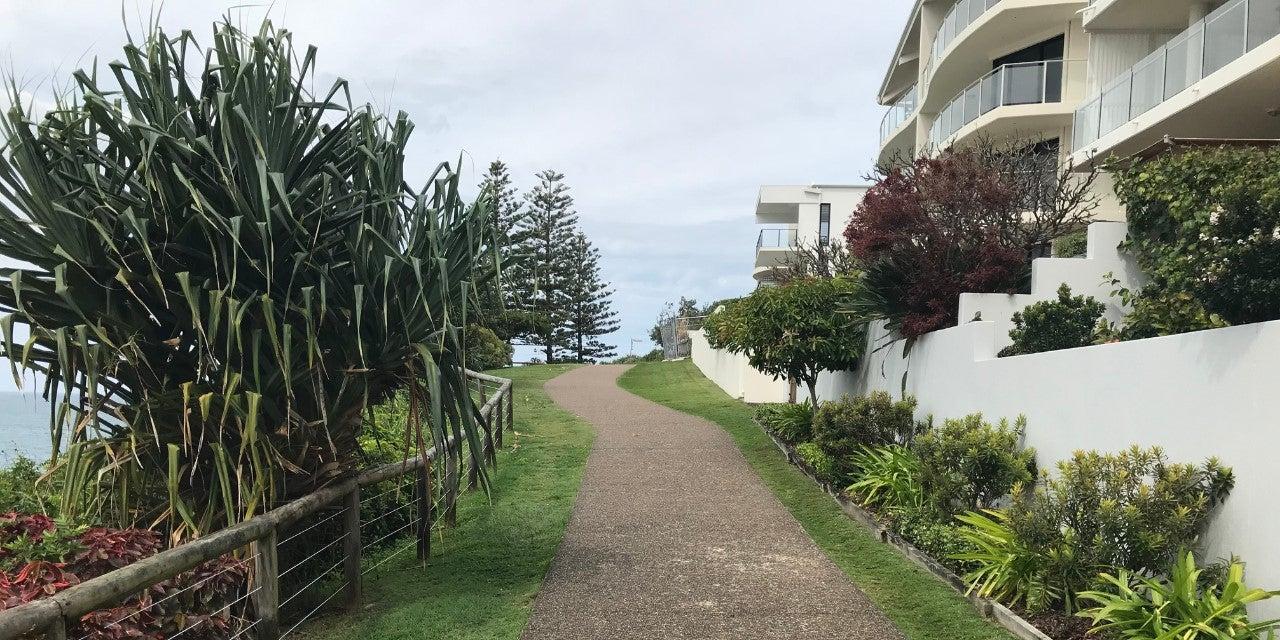 A scenic pathway on a headland, with lush trees lining the left side and modern apartments on the right, under a clear sky.