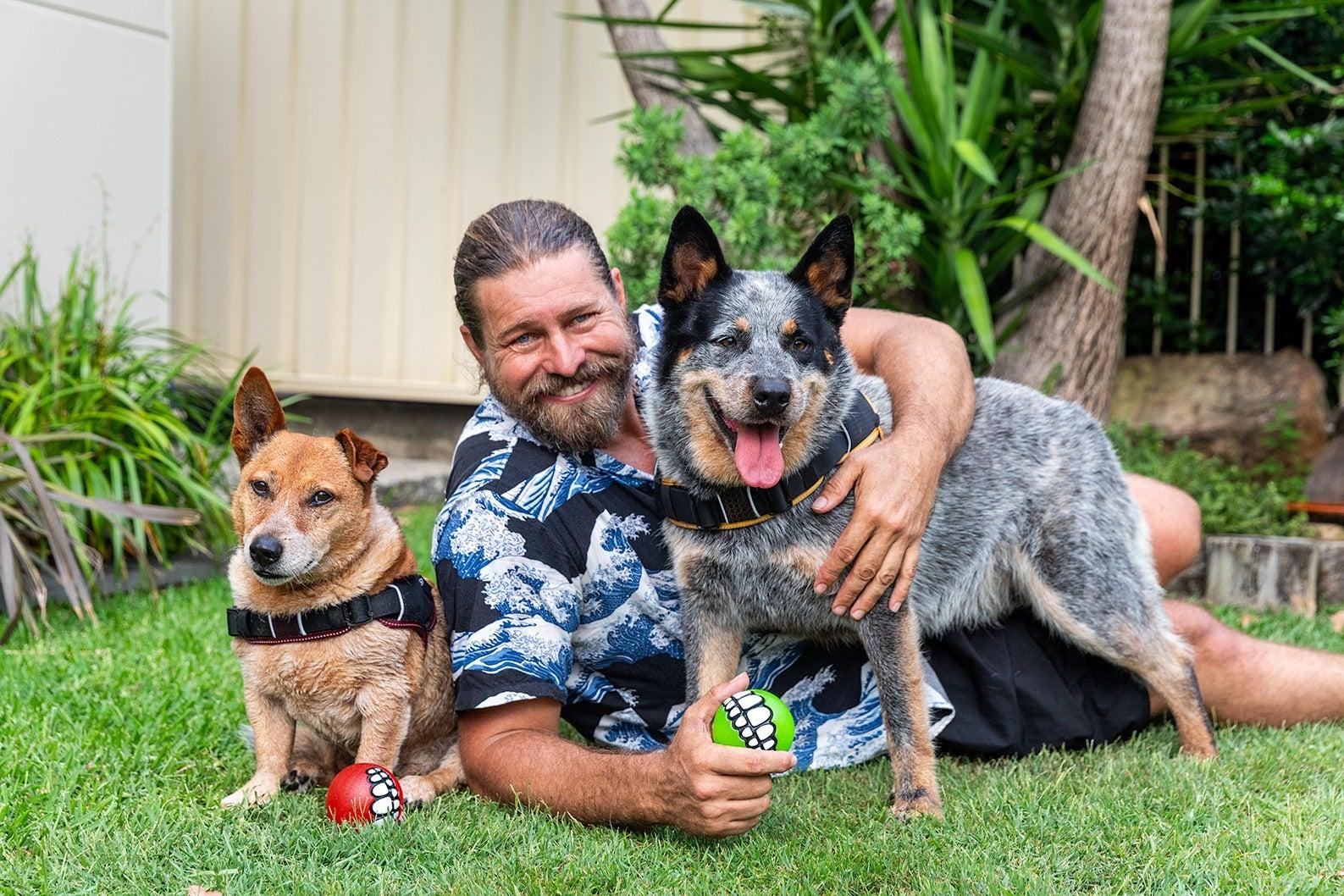 A man holding a ball, lying on the grass in between two dogs with his arm around one of the dogs