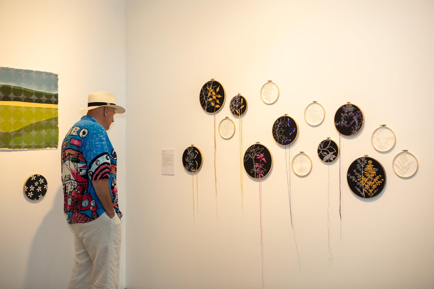 A man in a hat standing in front of a range of artworks at the Wildflowering by Design exhibition launch