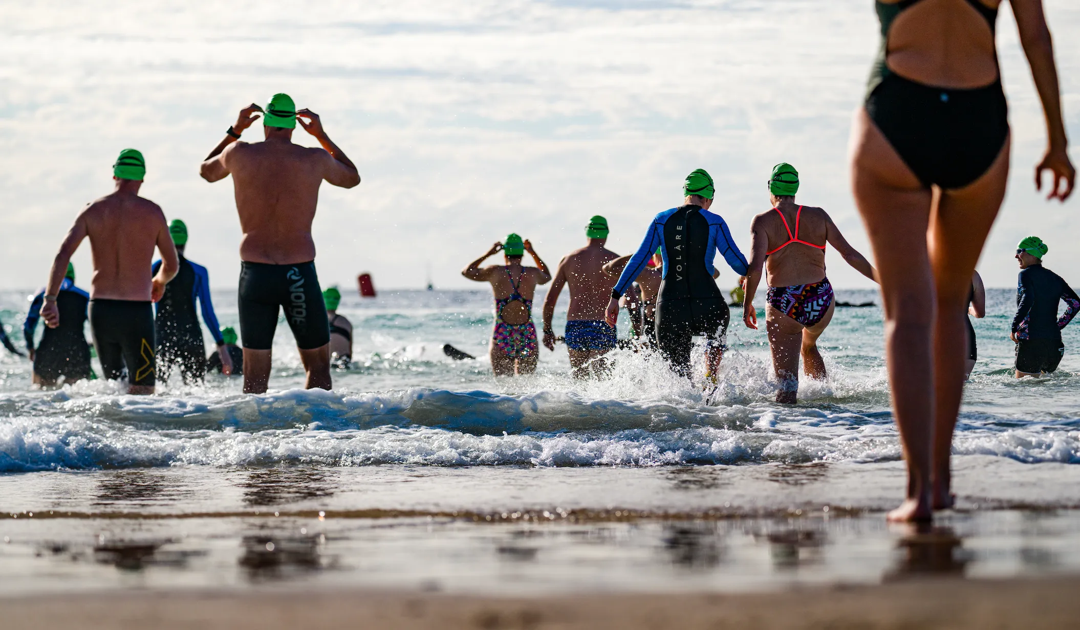 A large group of people in swimsuits and bathing caps are running into the ocean and swimming.