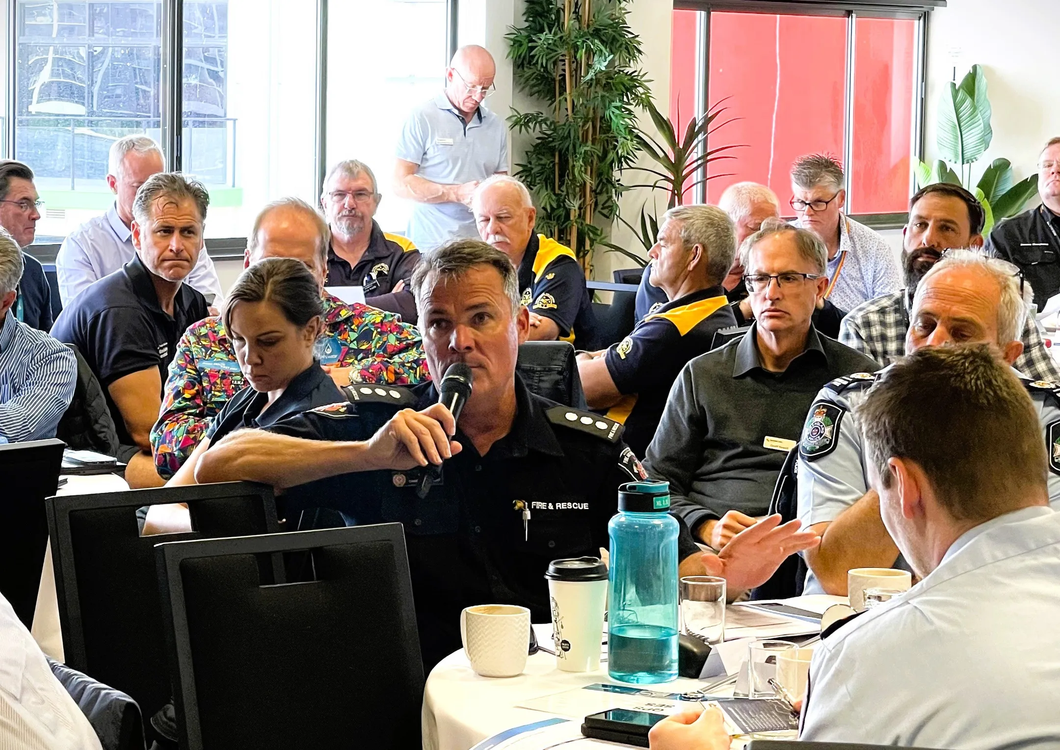 A room full of men and women from Emergency Services and Sunshine Coast Council, are seated at tables. The man from Fire and Rescue is speaking into a microphone. Everyone looks serious and attentive.