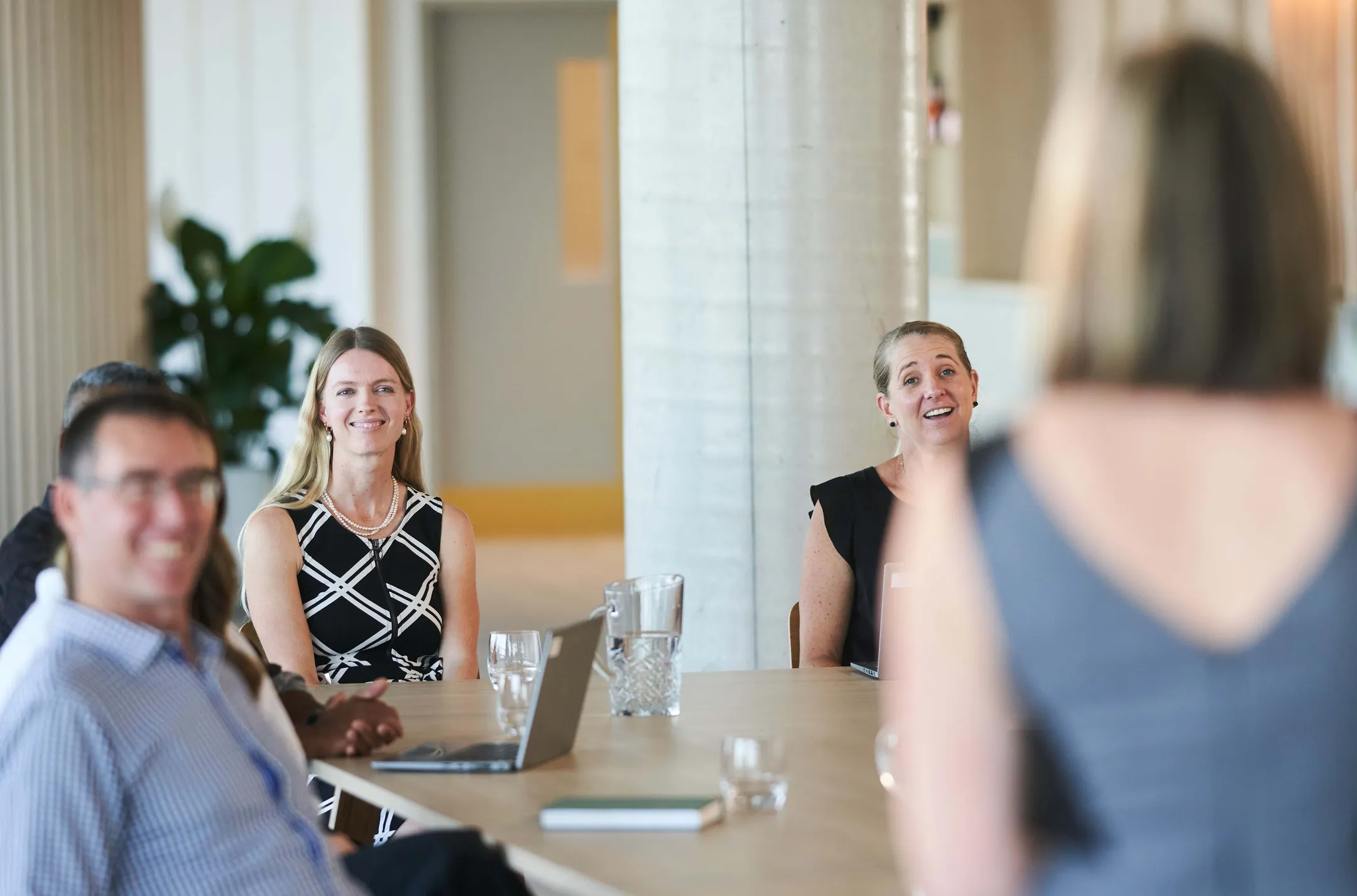 A young professional looking woman sit at a table with a group of other people. She smiles at someone off camera.