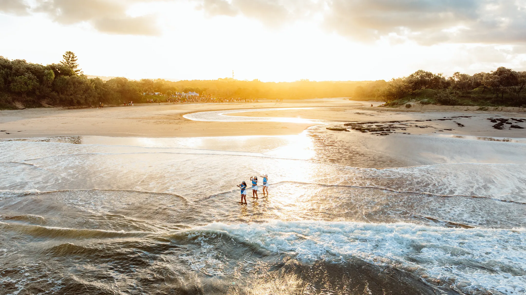 A landscape scene - looking from the ocean towards land, with small waves/white wash in the forefront, with three people standing in the shallows and a backdrop of sand, trees and golden sunlight.