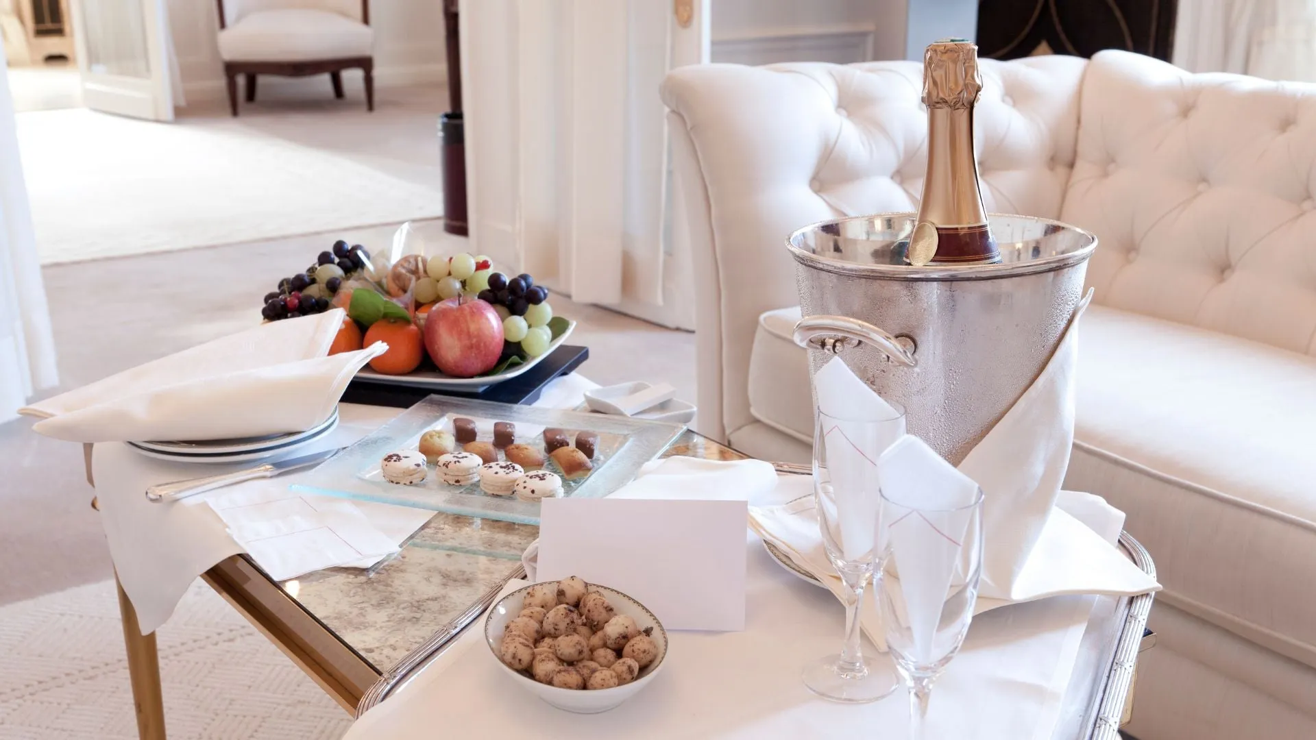 Luxury hotel room with white furnishings and decore. A table laiden with a platter of delicious and colourful fresh fruits of apples and grapes. A bottle of champagne chilling in a silver champagne bucket. Two champagne glasses with elegantly folded white napkins.
