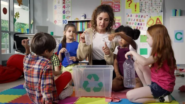 Teacher sitting on the floor in a classroom with small children around her looking at empty plastic bottles with a clear box with the recycling symbol in the middle of them all.