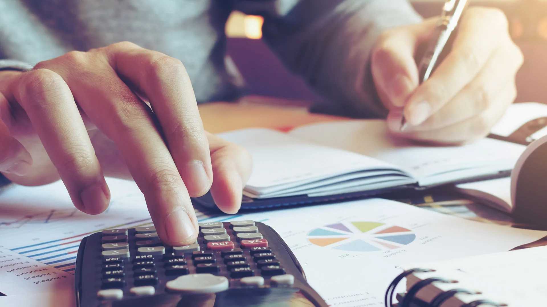 Person writing in book and using calculator with documents over desk.