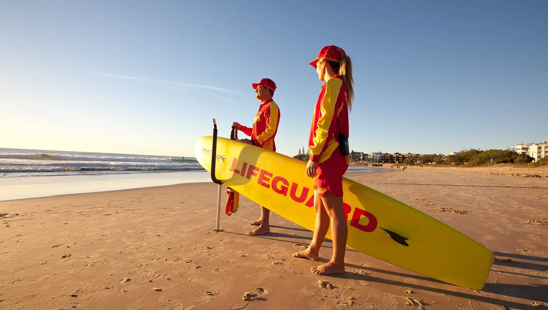 Two life guards looking out to sea from the beach with a rescue board between them.