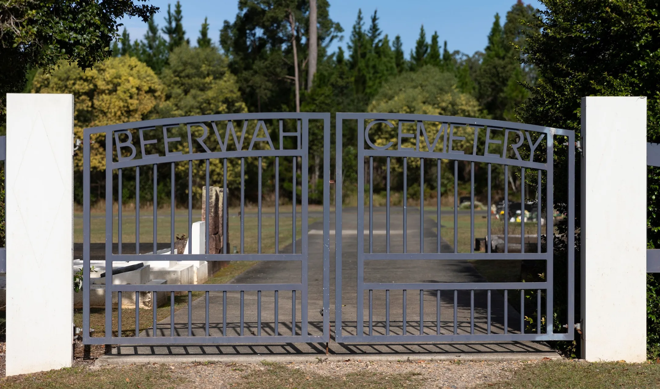 Closed entry gate with the words 'Beerwah Cemetery' forming part of the metal gate, two white pillars on either side and a footpath leading through the burial section to lawns and mature gardens beyond