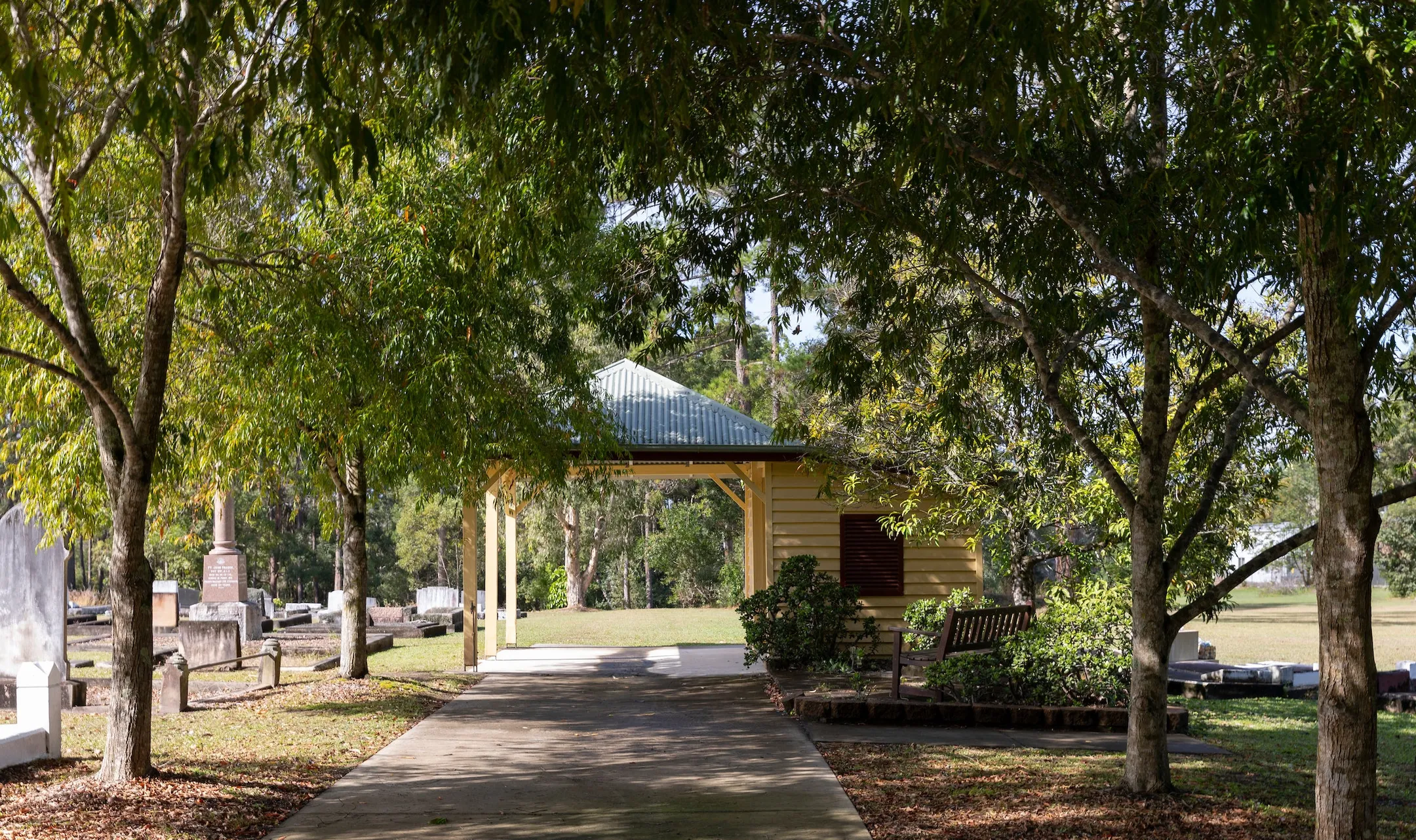 Tree lined wide concrete pathway to covered shelter - monumental section on left hand side and a shaded garden and seating area on the right side.