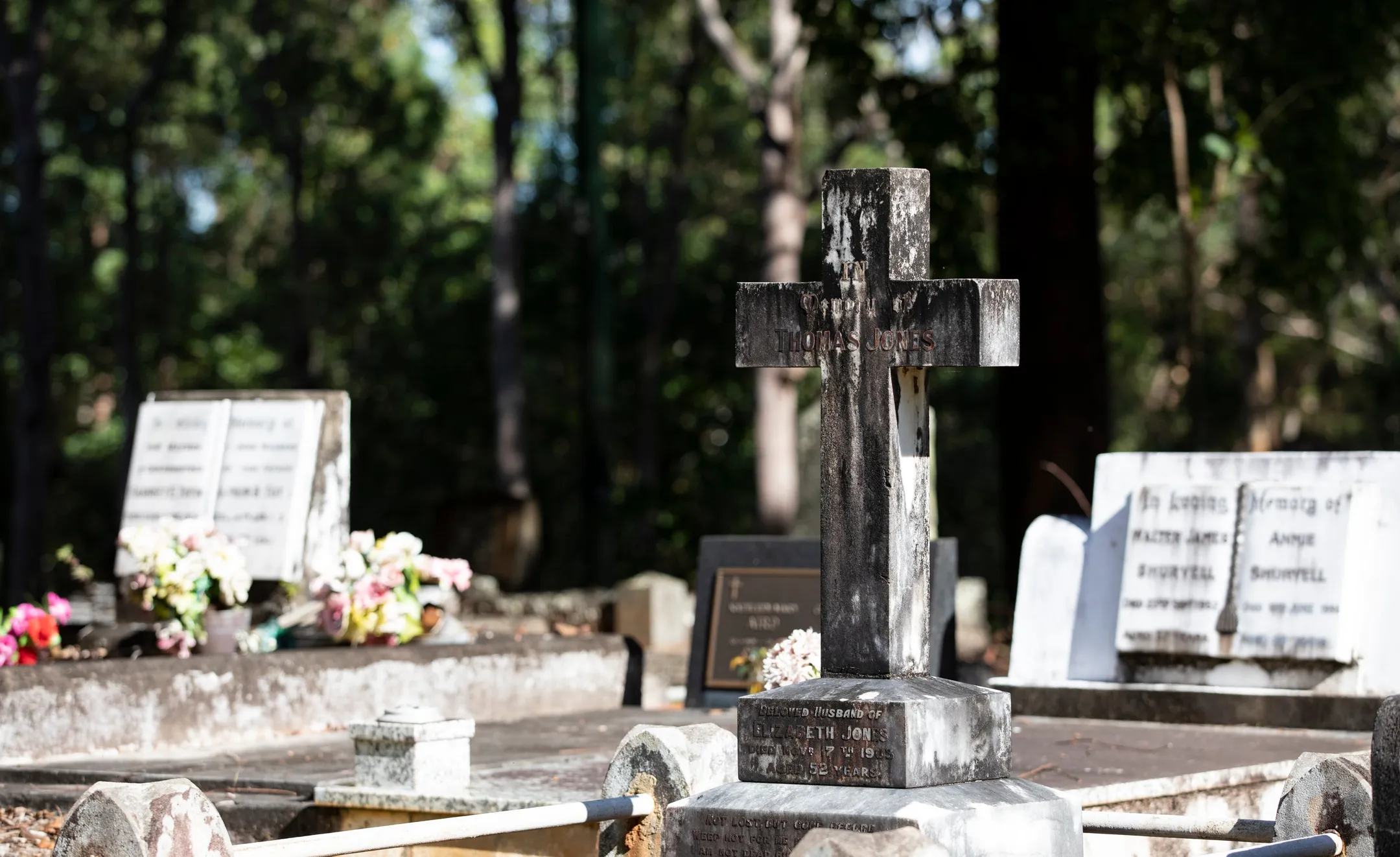 Old headstone in shape of a cross on a stone base in monumental section of cemetery.