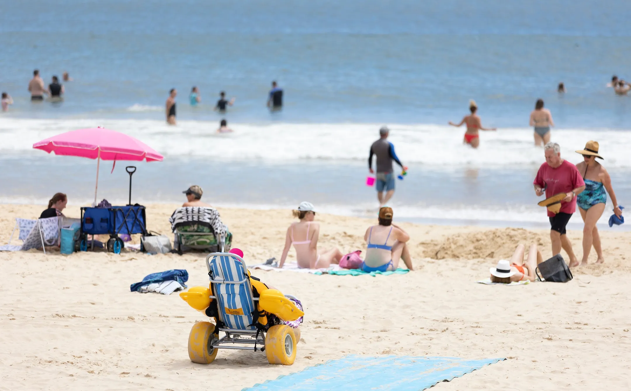 A accessible beach wheelchair and mat on the sand, surrounded by people sitting on the beach and swimming in the ocean.