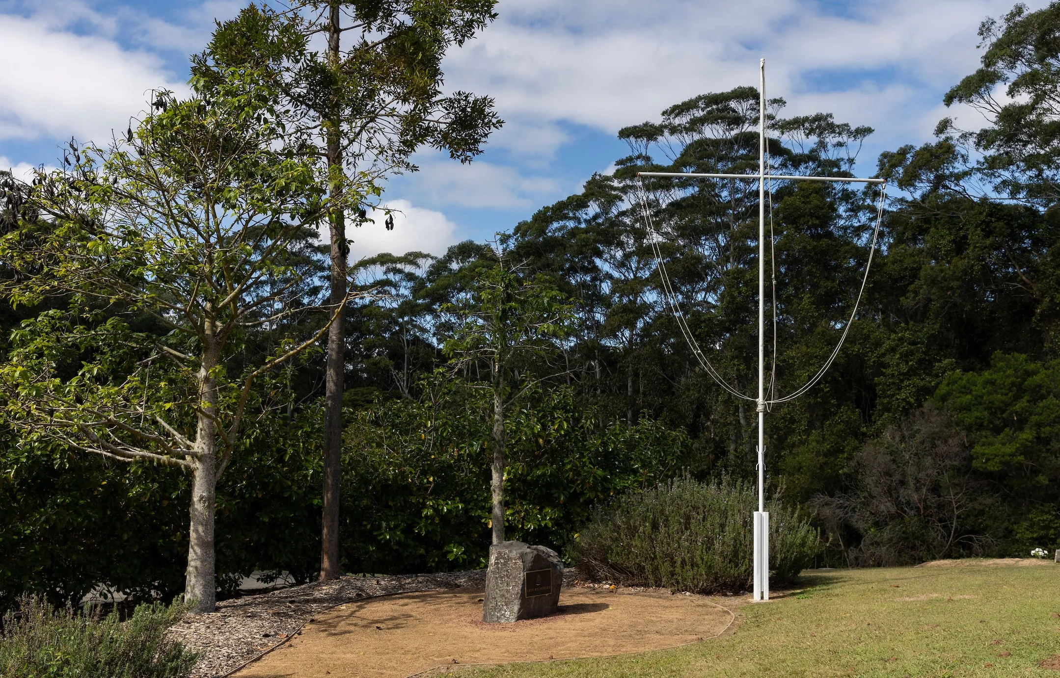 Cleared gravel section with a boulder in the centre and a memorial plaque, and mature trees and gardens in the background