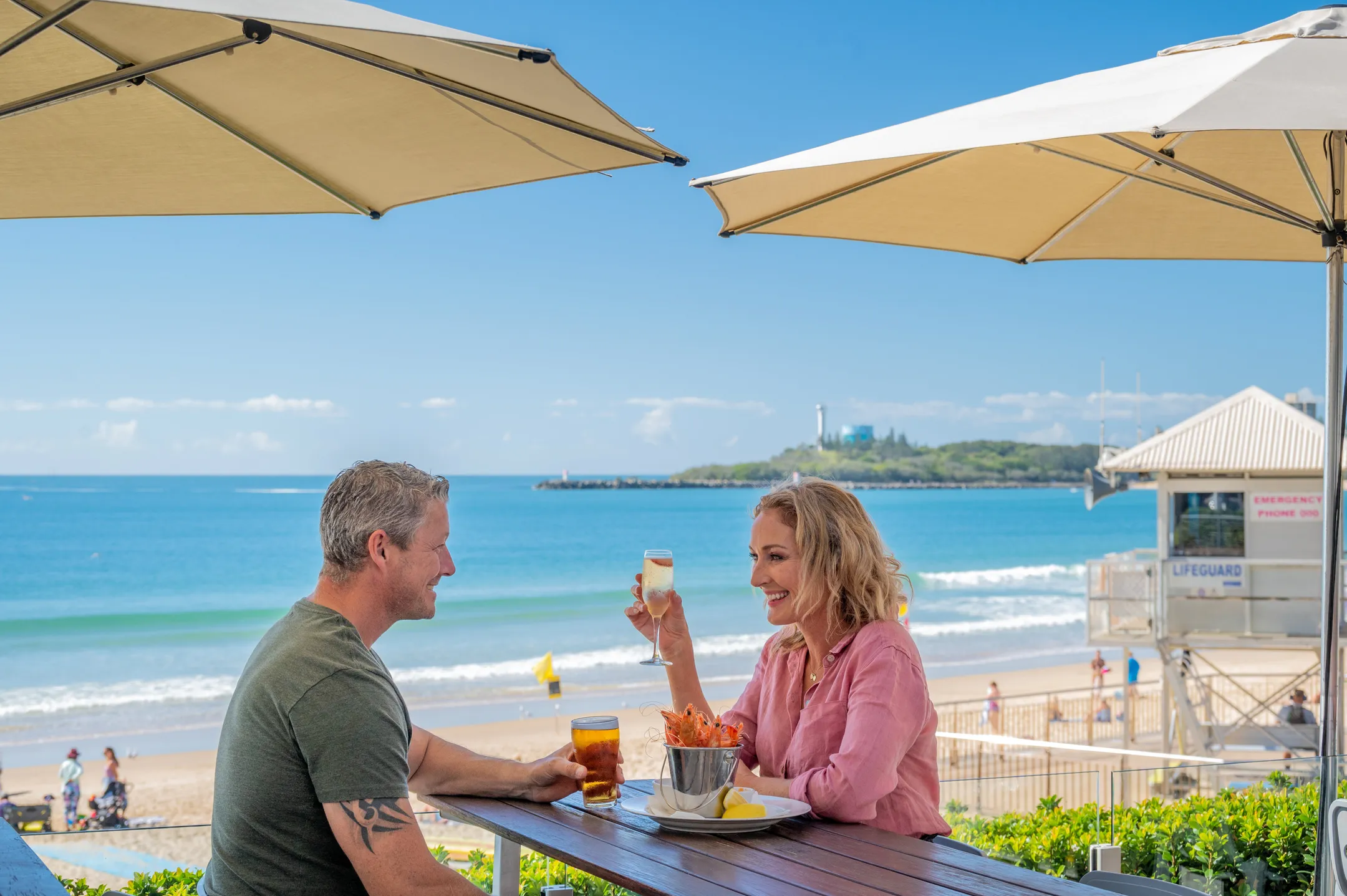 A couple eating and drinking on the deck of Mooloolaba Surf Club with the ocean in the background.