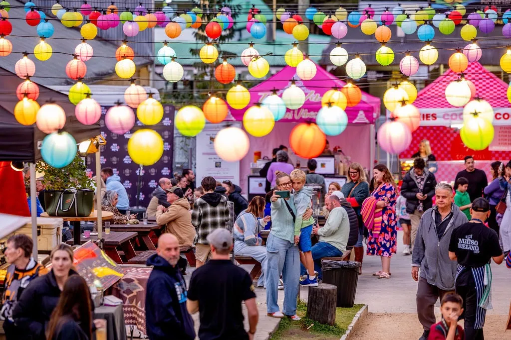 Colourful lantern balls hanging from wire above many people sitting and standing around - and in the background you can just see some colourful tents - food providers.