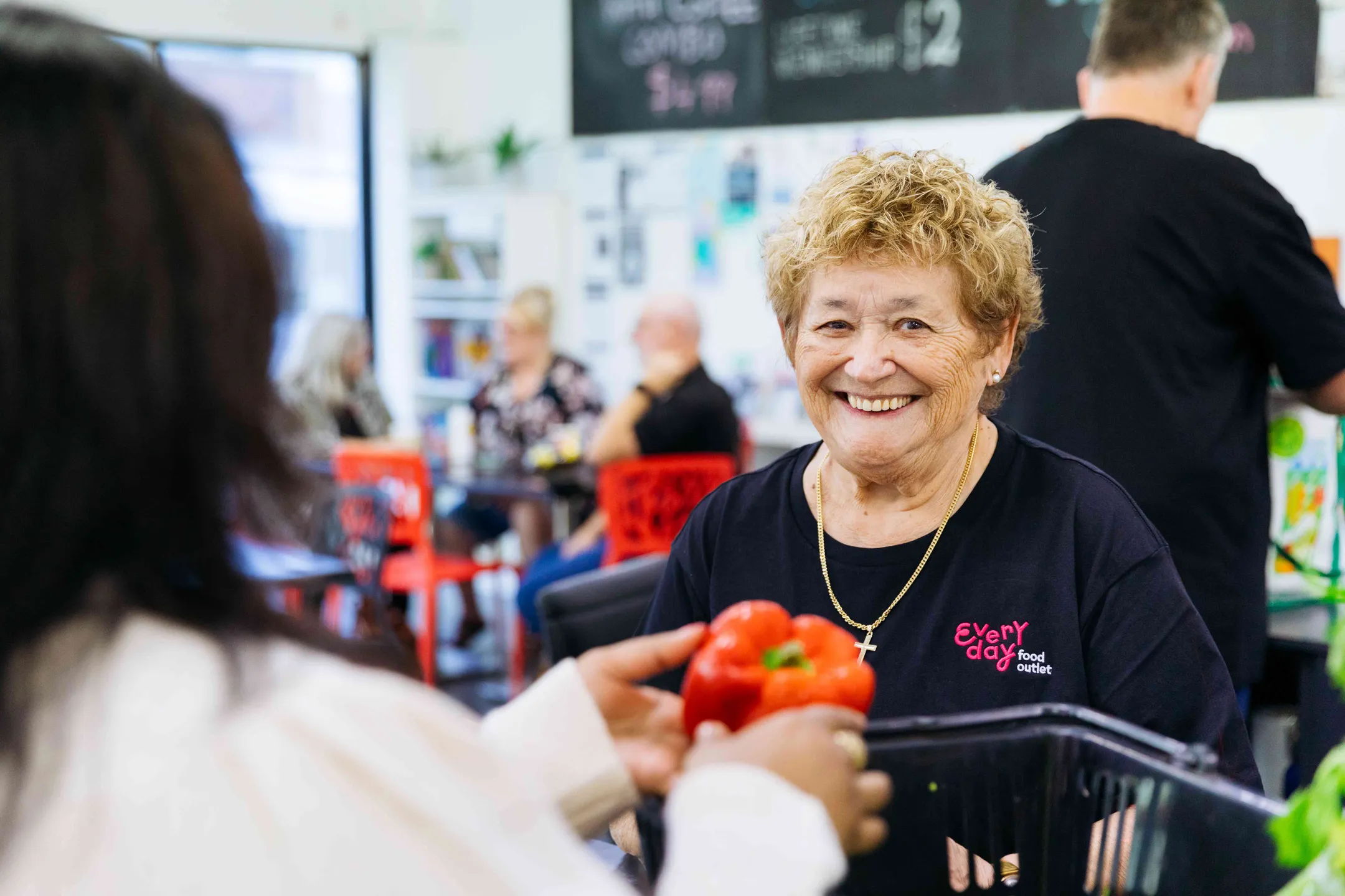 An older woman wearing a black shirt with a logo, smiles at the camera. The woman in front of her is washing a capsicum.
There is a group of people also wearing black shirts, in the distance talking.