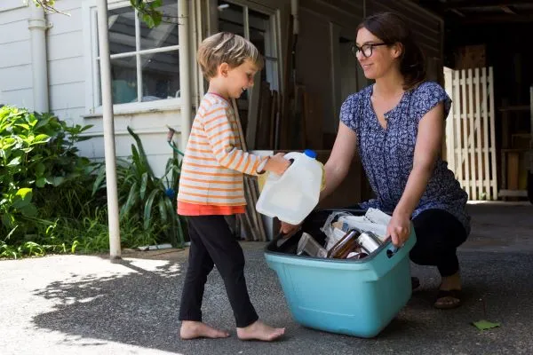 Lady holding a recycling tub for a little boy to put a empty plastic bottle into it.