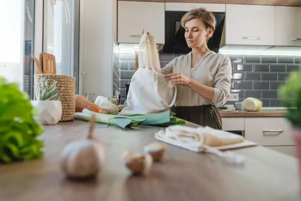 A lady in her kitchen examines the groceries she brought home in a reusable bag.