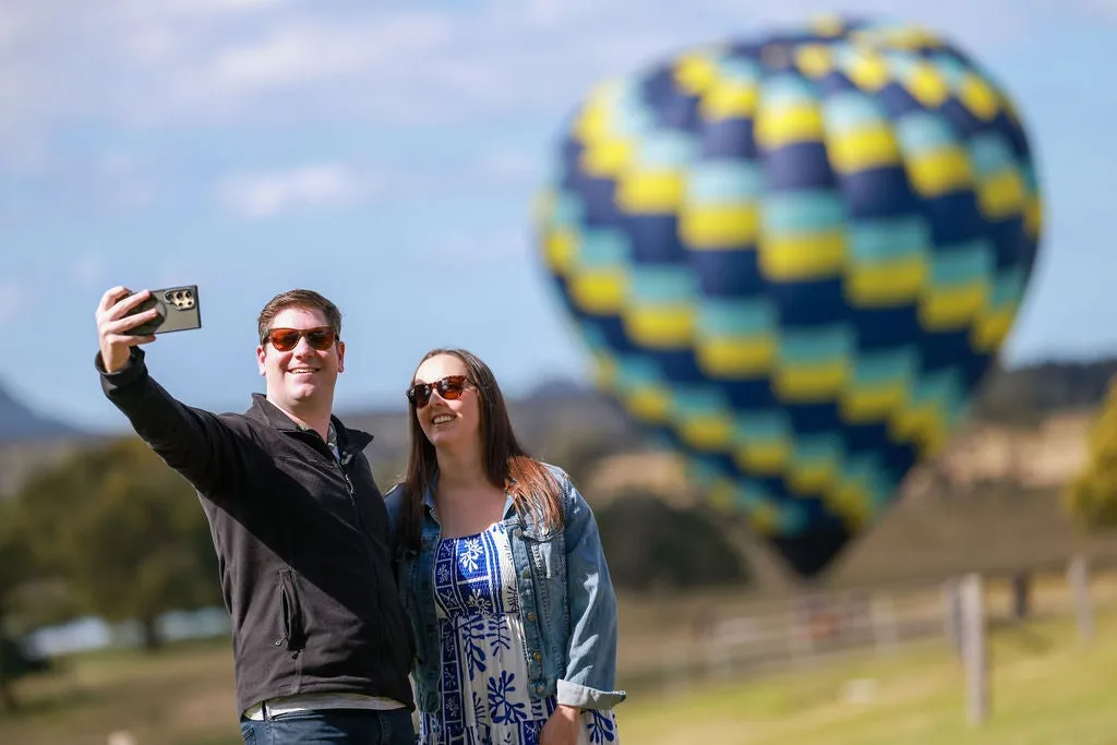 Two people holding up a phone/camera to take a selfie with a blue and yellow hot air balloon blurred in the background behind them.