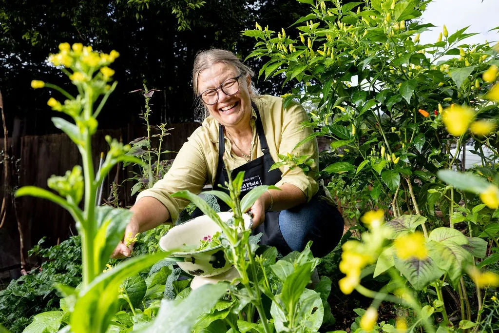 A lady holding a bowl in the middle of a green garden with some yellow flowers here and there. She is collecting plants for cooking.