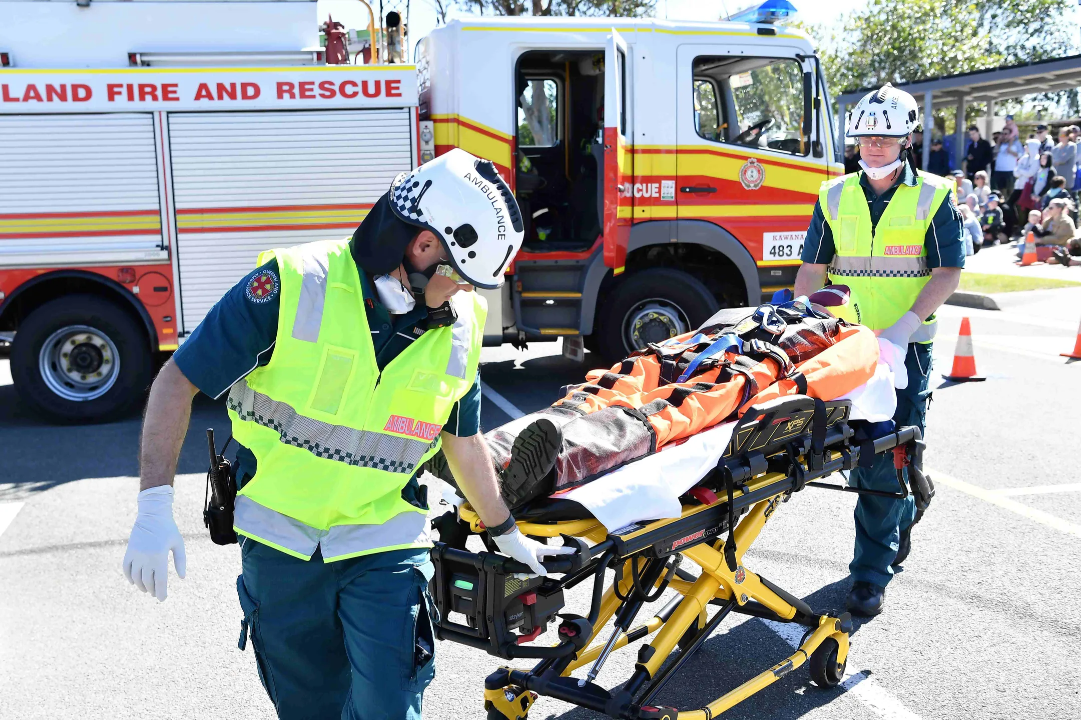 Two Ambulance officers enact an emergency rescue with a mannequin on a stretcher. IN the background is a fire truck and a crowd of onlookers.