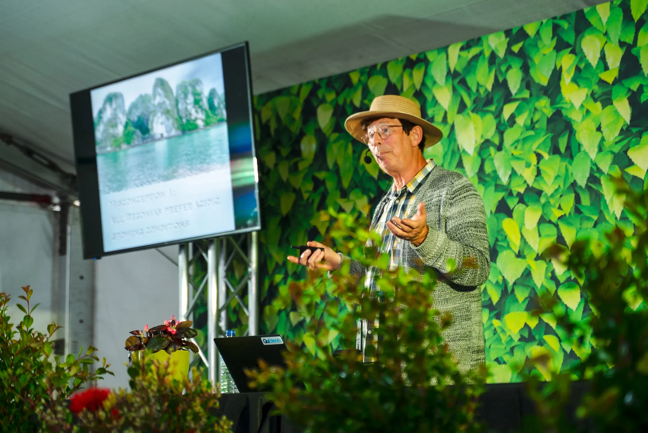 ABC Gardening Australia personality Jerry Coleby-Williams standing on stage (he is partially covered by some plant form in the forefront of the picture) with a digital green leaf background behind him and a TV on the left to support his presentation.