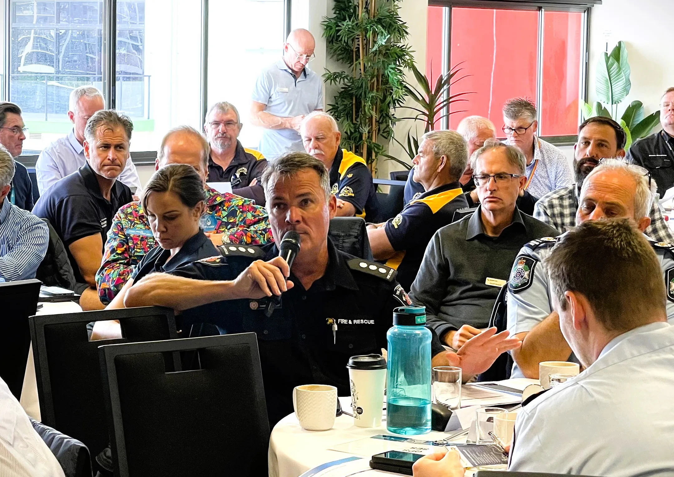 A room full of men and women from Emergency Services and Sunshine Coast Council, are seated at tables. The man from Fire and Rescue is speaking into a microphone. Everyone looks serious and attentive.