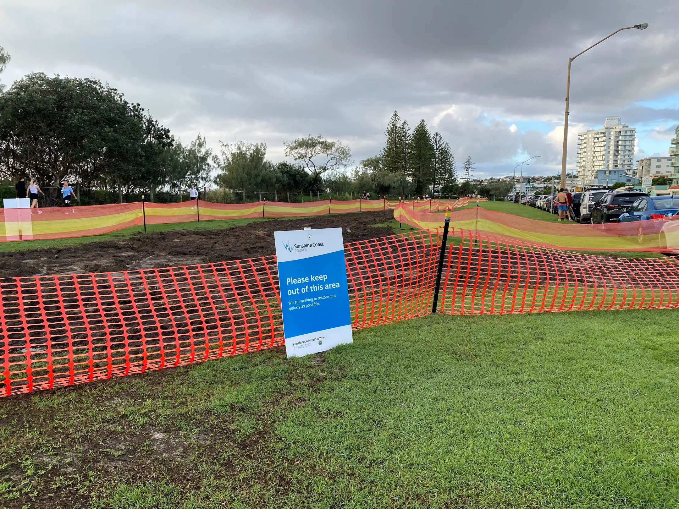 Muddy and damaged parkland turf at Alexandra Headland, surrounded by an orange fence and Council sign that says please keep out of this area.