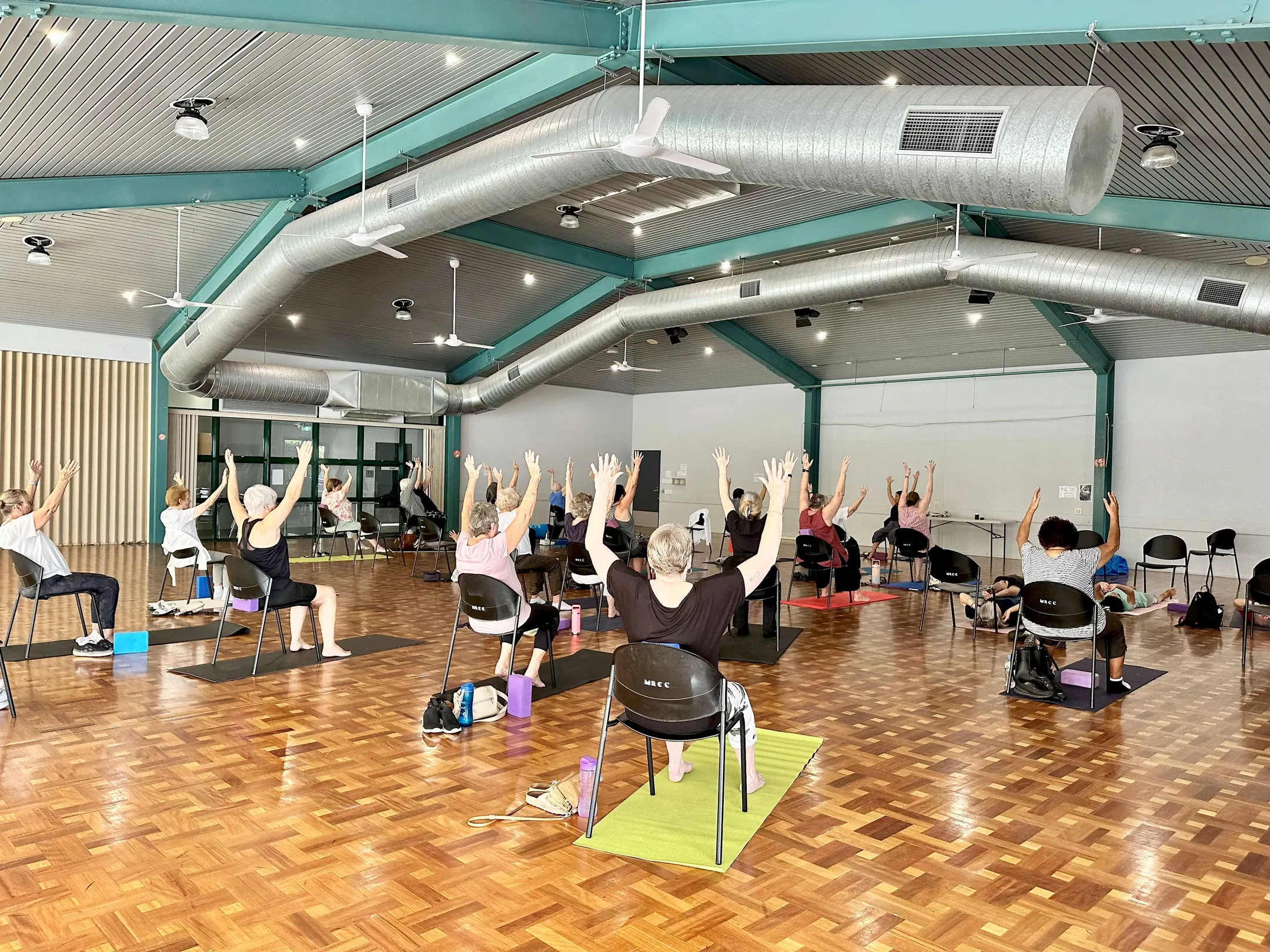 Group of people doing Healthy Sunshine Coast Chair Yoga enjoying health and wellbeing exercises. Featured in a hall.