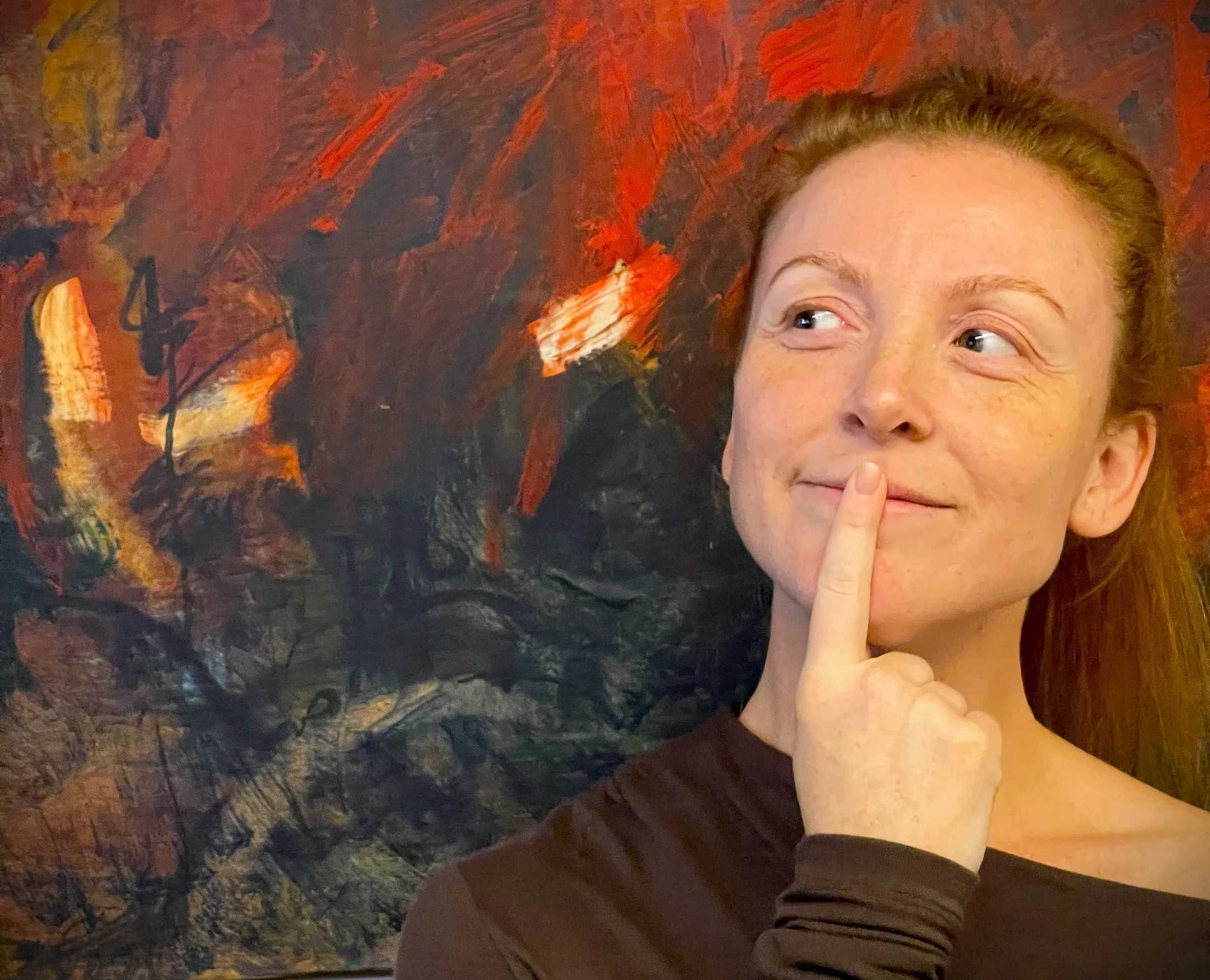 A young, brown eyed, red headed woman stands in front of a red and brown abstract painting with a pointer finger on her mouth and looking cheekily to the side.