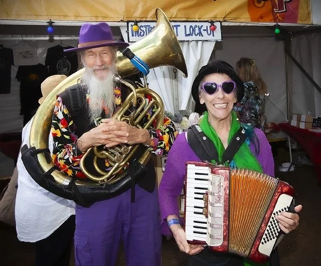 A smiling, grey haired man and woman are wearing vibrant clothing and hats. The man has a tuba and the woman has an accordion. The duo are standing in front of a festival tent.