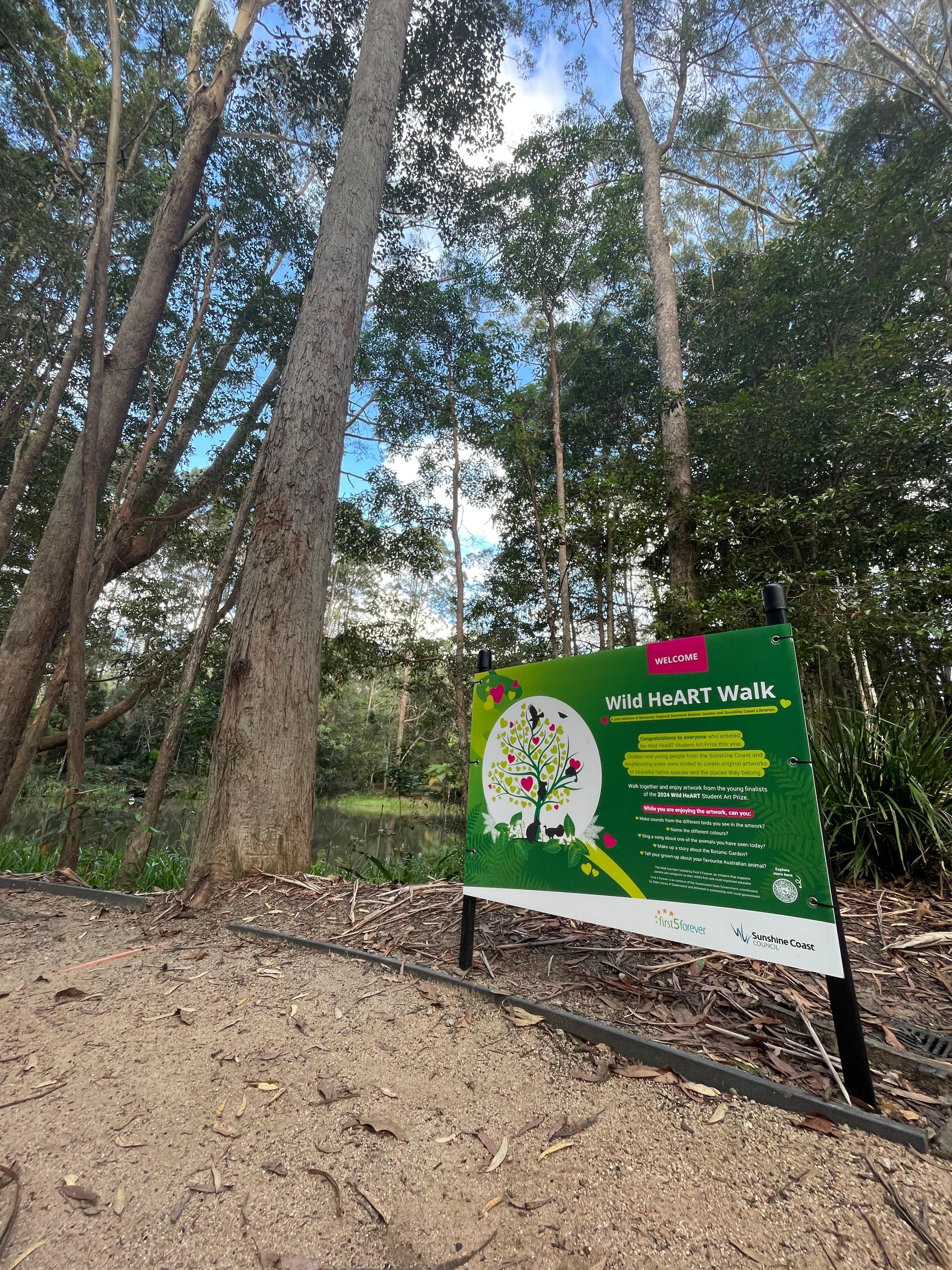 Forest with tall trees with green sign in the foreground. A walking trail in the front of the image.