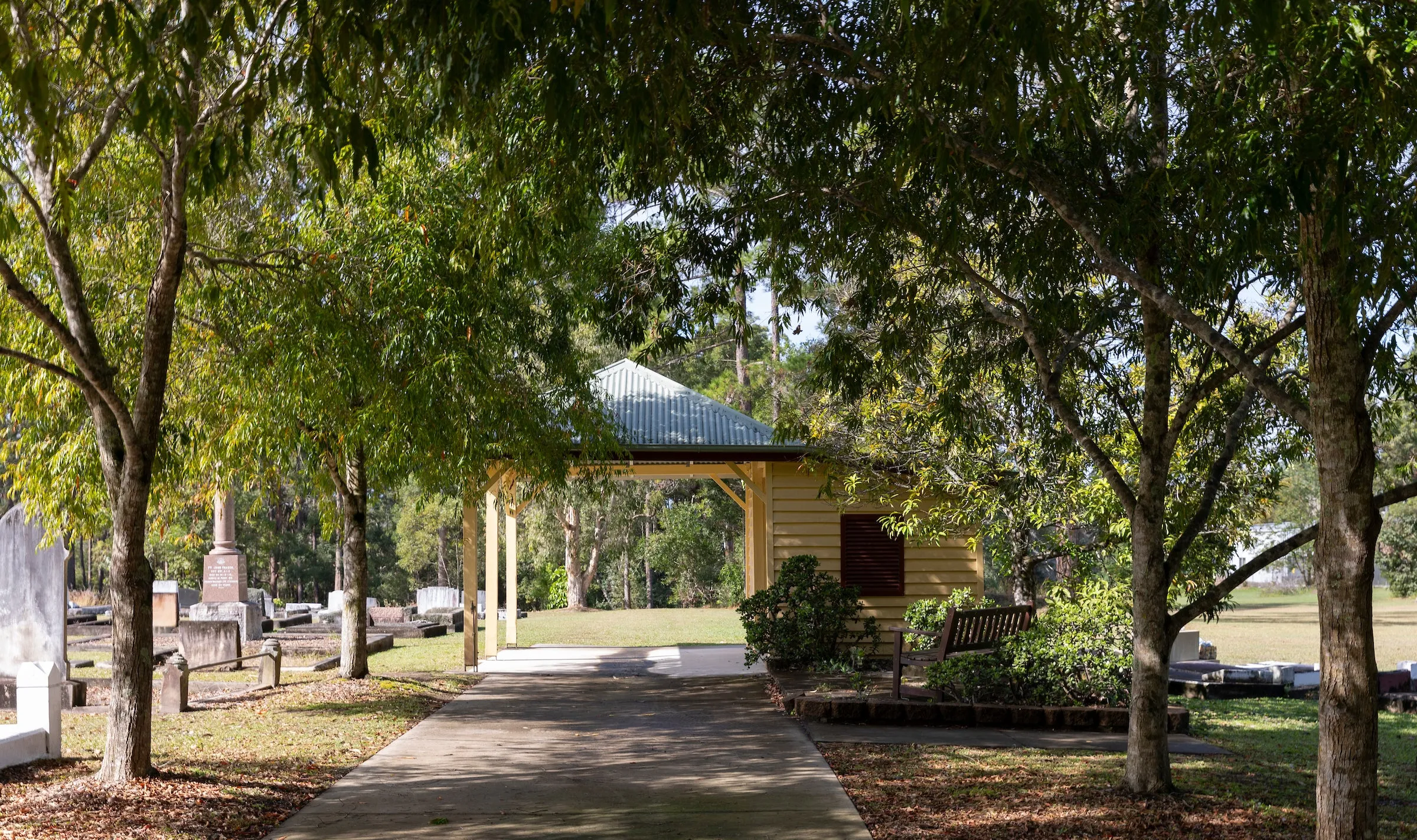 Tree lined wide concrete pathway to covered shelter - monumental section on left hand side and a shaded garden and seating area on the right side.