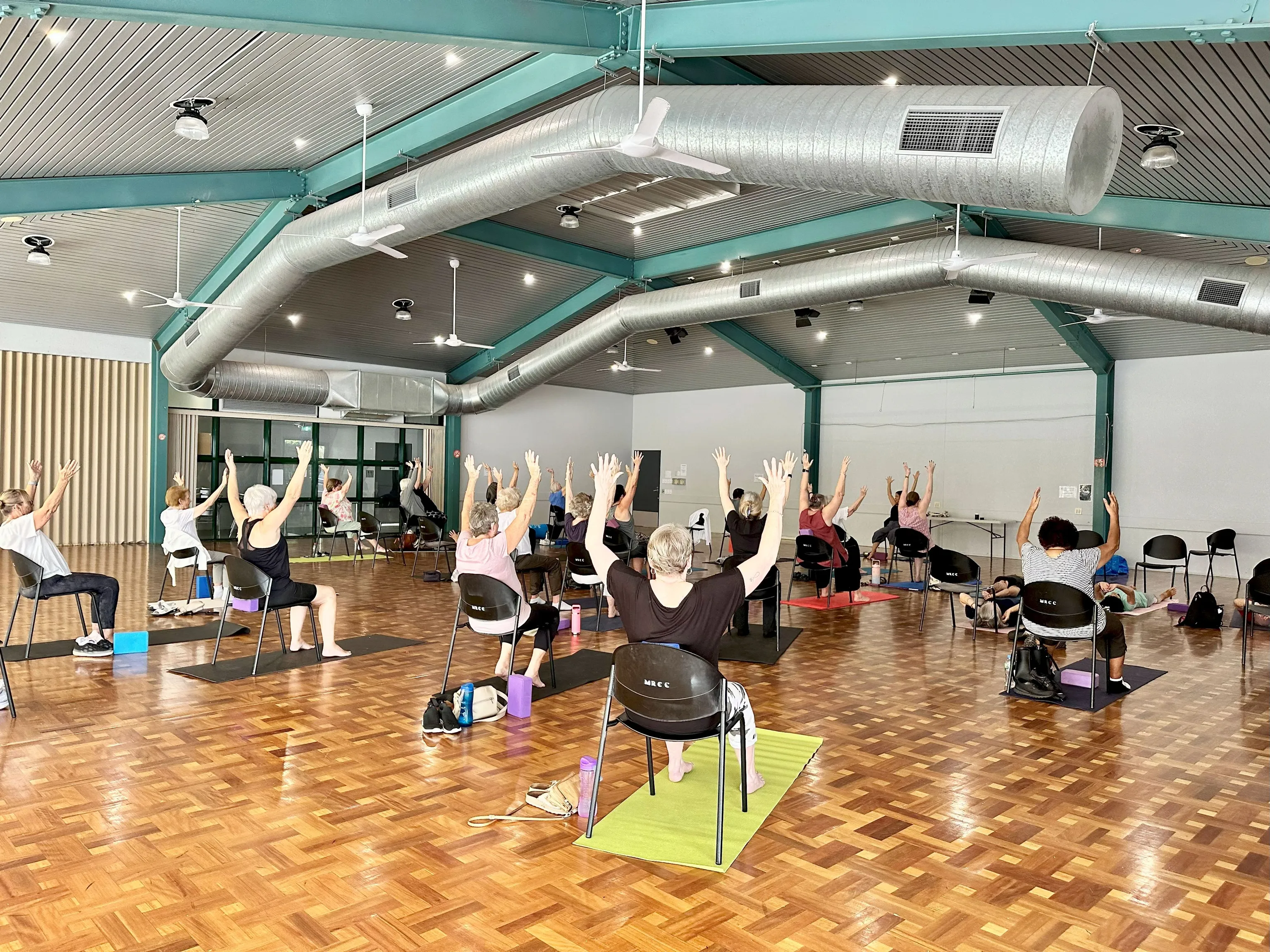 Group of people doing Healthy Sunshine Coast Chair Yoga enjoying health and wellbeing exercises. Featured in a hall.