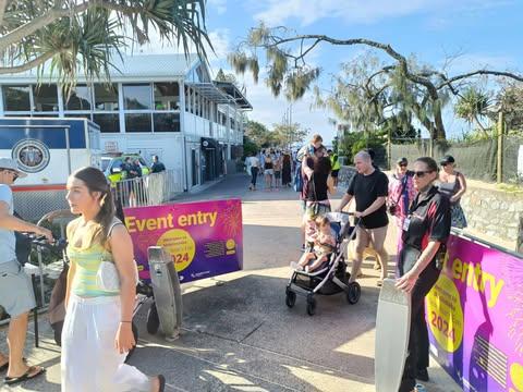 Pink, purple and yellow event entry signage, security person on the right, and patrons (including one with a pram) entering/exiting the site at Mooloolaba - with the Mooloolaba Surf Club building in the background on the left.