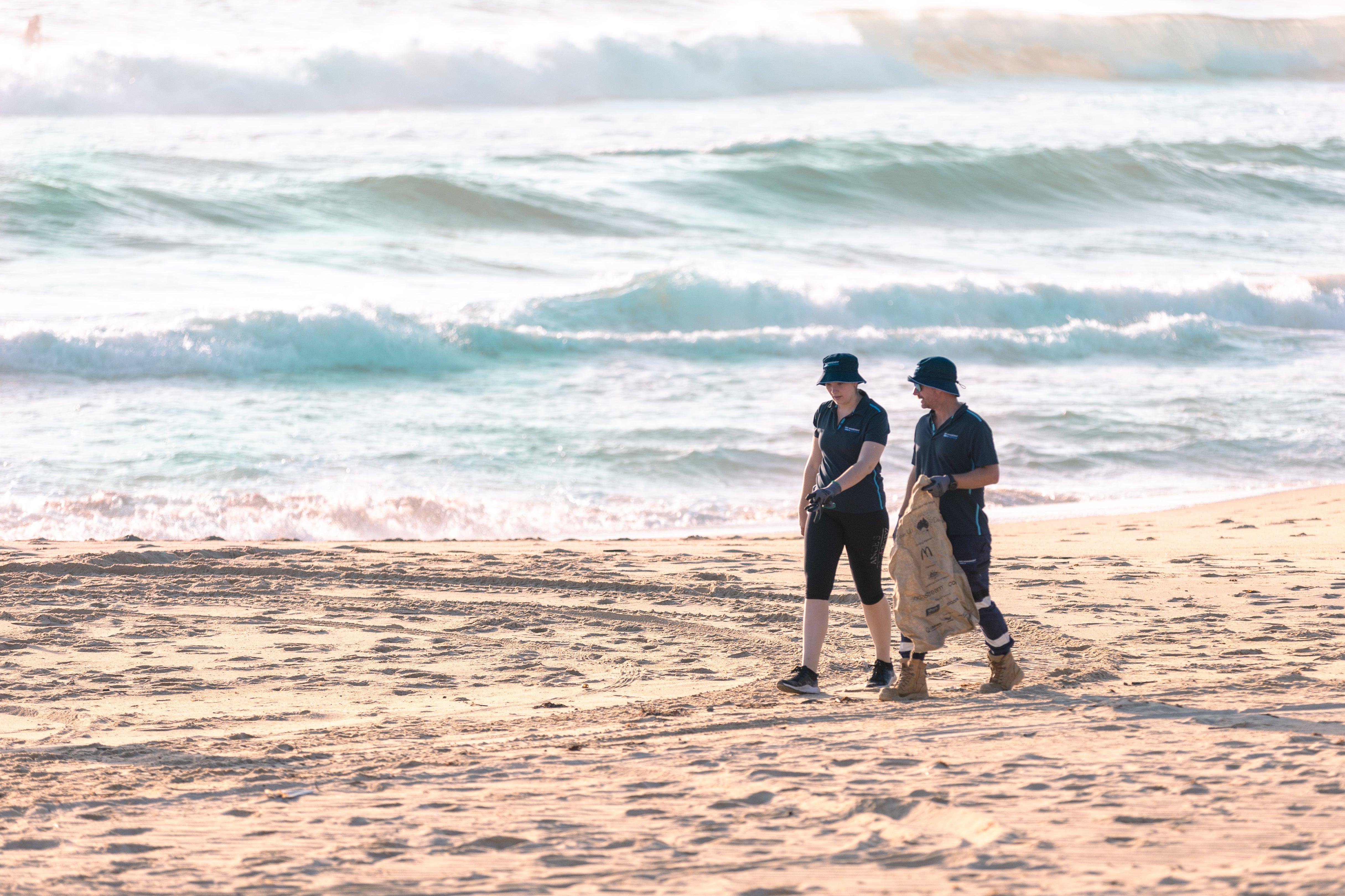 A group of people collecting rubbish on a sandy beach, working together to clean up the environment. They have a white bag in their hand to contain the rubbish.