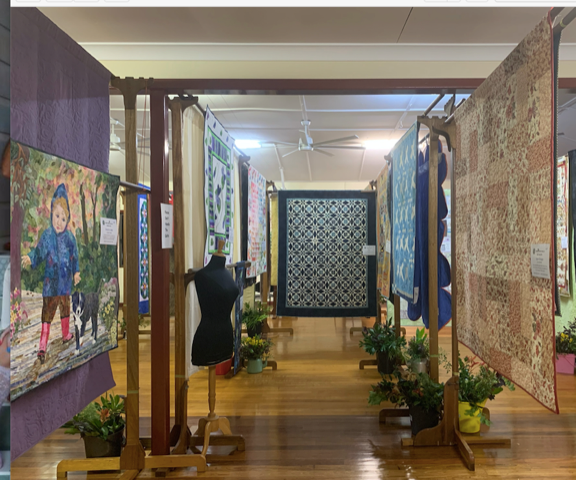 Wooden floors in a room full of colourful artwork and paterned rugs on display on poles. Green lush plants on display.