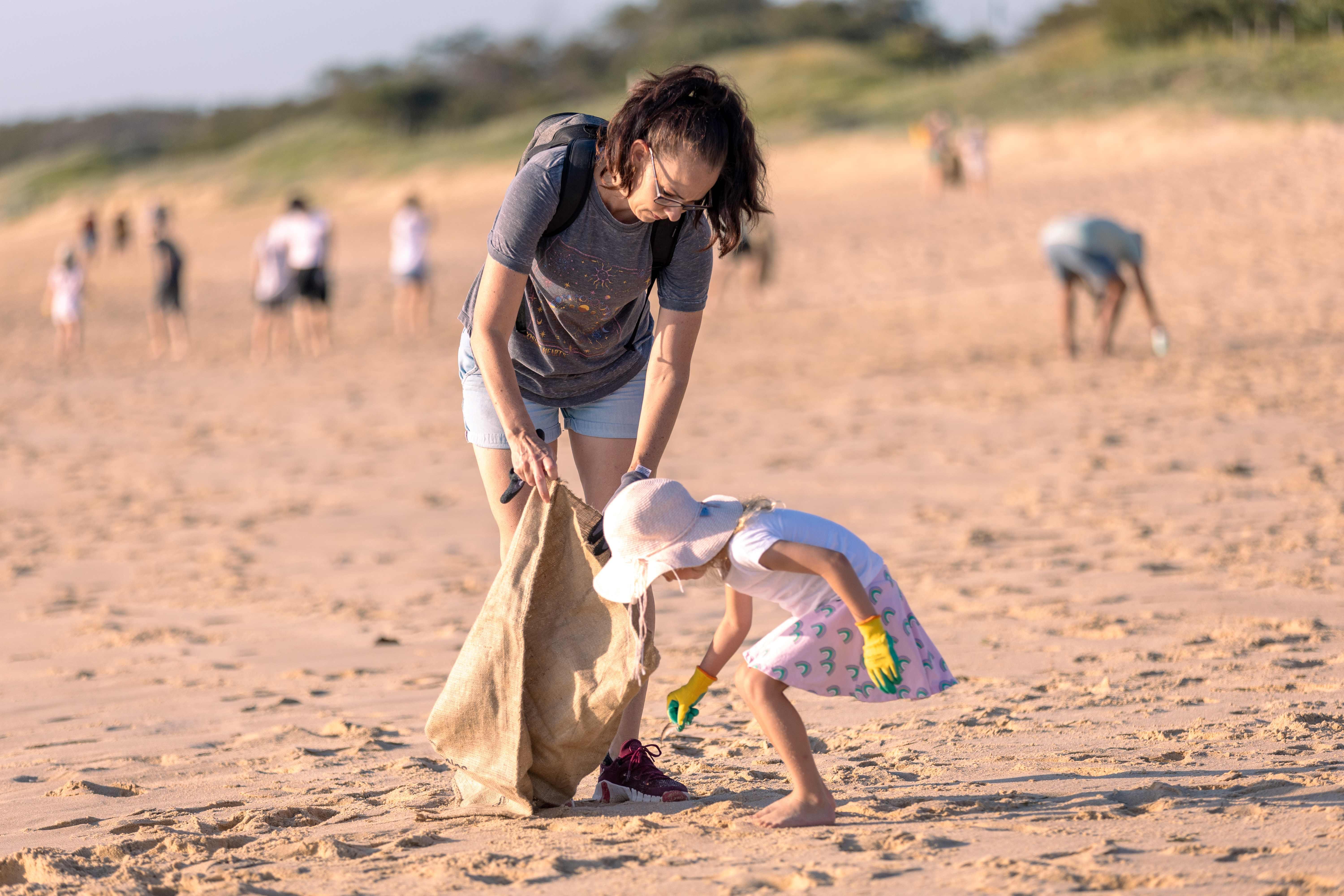 A adult female and young child collecting rubbish on a sandy beach, working together to clean up the environment.