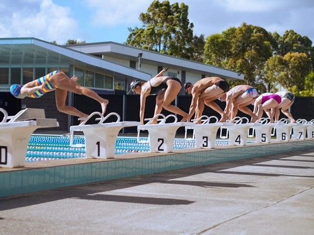 Six professional swimmers on starting blocks around an outdoor swimming pool jumping in to start a race.